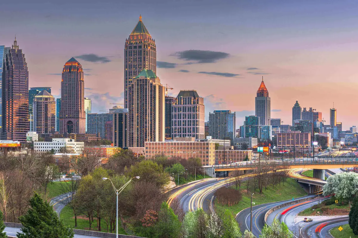 Atlanta_Skyline_Abendverkehr.jpg Skyline von Atlanta, USA, bei Sonnenuntergang mit beleuchteten Hochhäusern und fließendem Abendverkehr auf den geschwungenen Highways im Vordergrund.