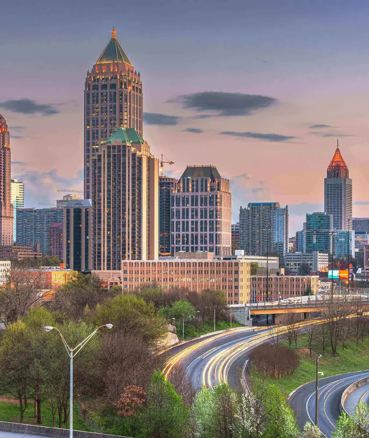 Atlanta_Skyline_Abendverkehr.jpg Skyline von Atlanta, USA, bei Sonnenuntergang mit beleuchteten Hochhäusern und fließendem Abendverkehr auf den geschwungenen Highways im Vordergrund.