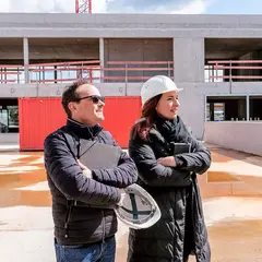 emmendingen-gebaeude-baustellenbegehung-2021.jpg Zwei Personen mit Helm und Unterlagen bei der Baustellenbegehung des neuen Firmengebäudes der Sutter Medizintechnik GmbH in Emmendingen. Im Hintergrund ist der Rohbau mit Betonfassade zu sehen.