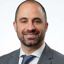 Porträtaufnahme einer Person mit kurzem Haar und Bart in grauem Sakko, weißem Hemd und Krawatte vor weißem Hintergrund