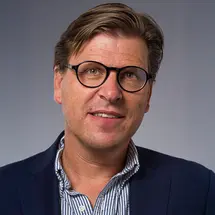 Dr. Meling mit Brille, dunkelblauem Sakko und gestreiftem Hemd vor grauem Hintergrund.