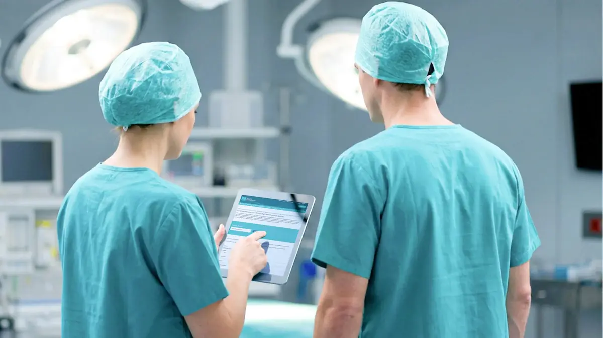 Zwei medizinische Fachpersonen in OP‑Kleidung betrachten gemeinsam ein Tablet mit einer digitalen Anwendung im Operationssaal.