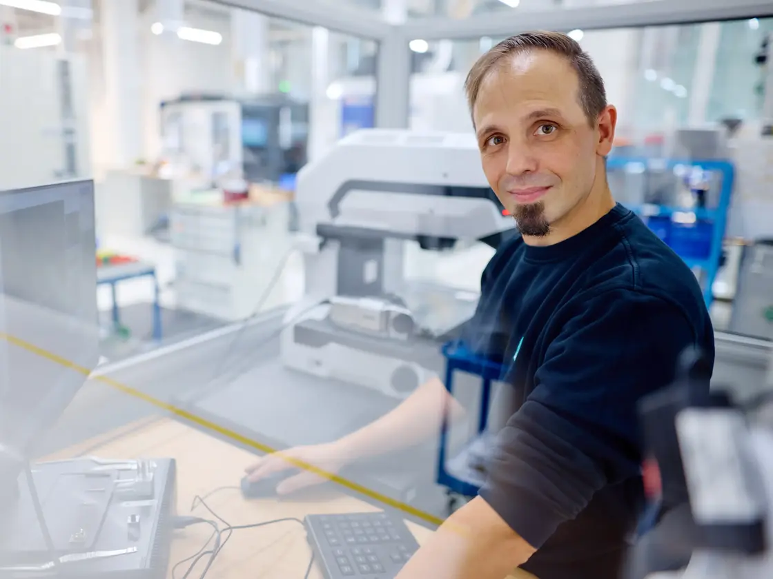 © Jörg Wilhelm Mitarbeiter von Sutter Medizintechnik steht an einem Arbeitsplatz in der CNC und blickt lächelnd in die Kamera. Im Hintergrund sind moderne Prüfgeräte und Fertigungsbereiche zu sehen.