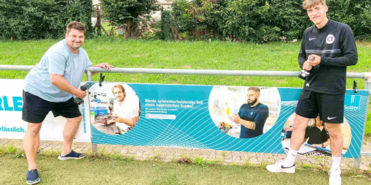 Zwei Männer montieren eine Sutter-Werbebande mit Karriereaufruf am Fußballplatz des FC Emmendingen, beide halten einen Akkuschrauber in der Hand und lächeln.